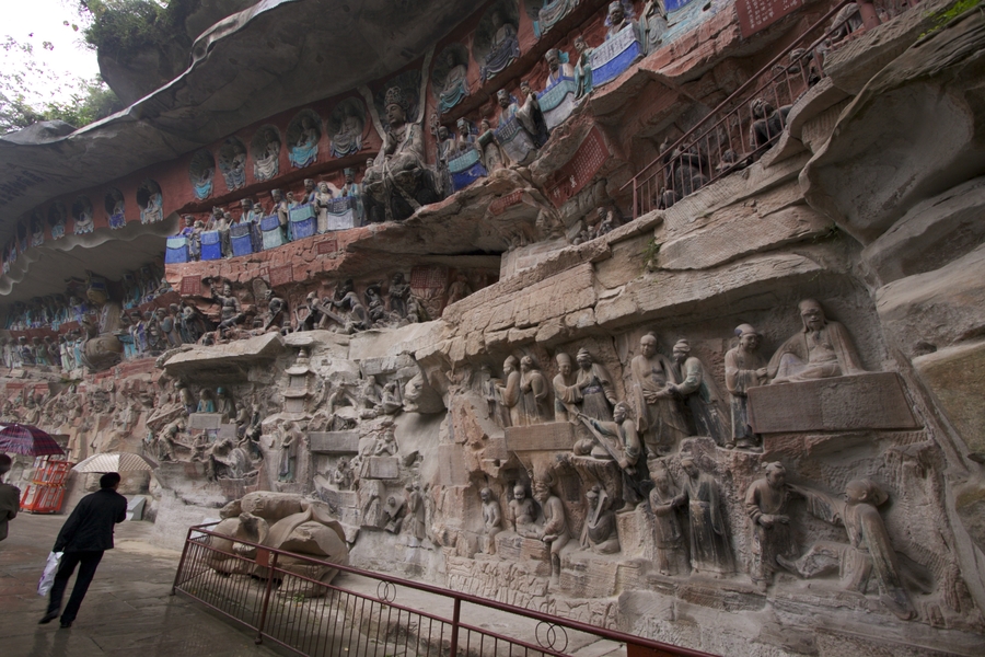 An immense rocky wall is carved all over with a series of sculptural groups in high relief. The upper register of figures includes a central buddha among seated attendants. This register is painted in blue and green against a red background. 