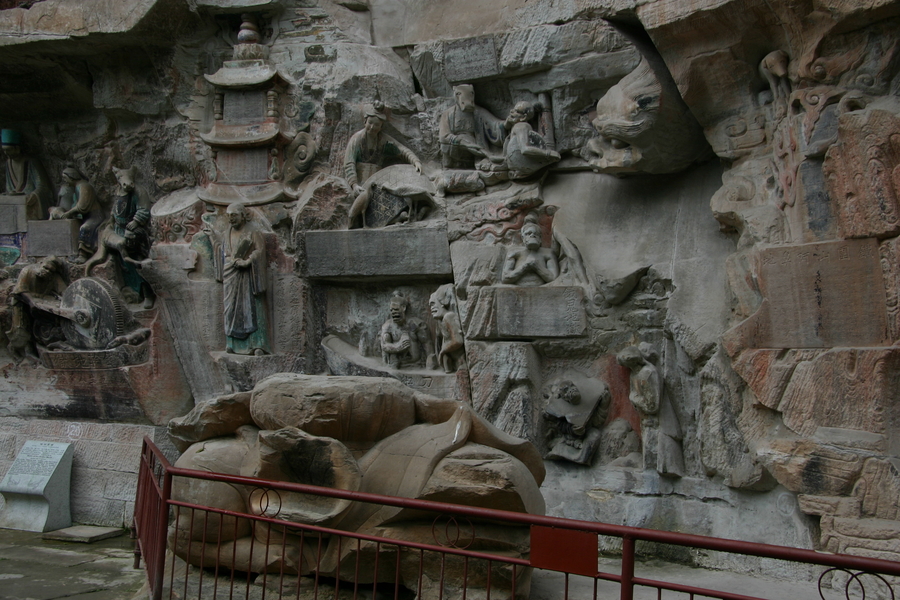 A stone, oblong altar stands in front of a rocky wall. Its curving form is abstract and vegetal. Behind the altar stands a rocky wall carved into a facade of high-relief stone sculpture.