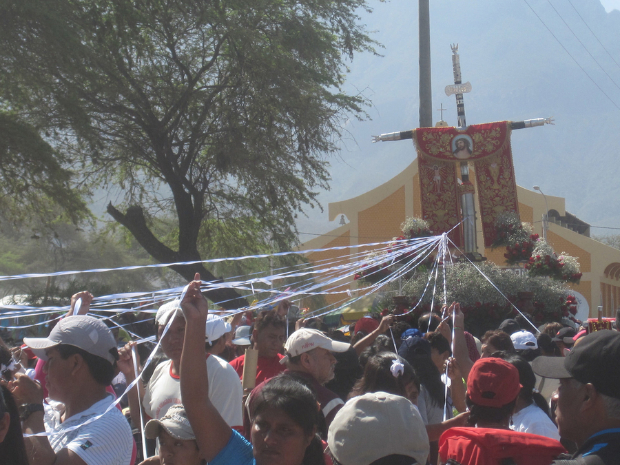 A group of tan-skinned pilgrims hold streamers that connect them to a processing cross. The wooden cross is decorated with a red and gold embroidered stole that bears an image of Christ. 