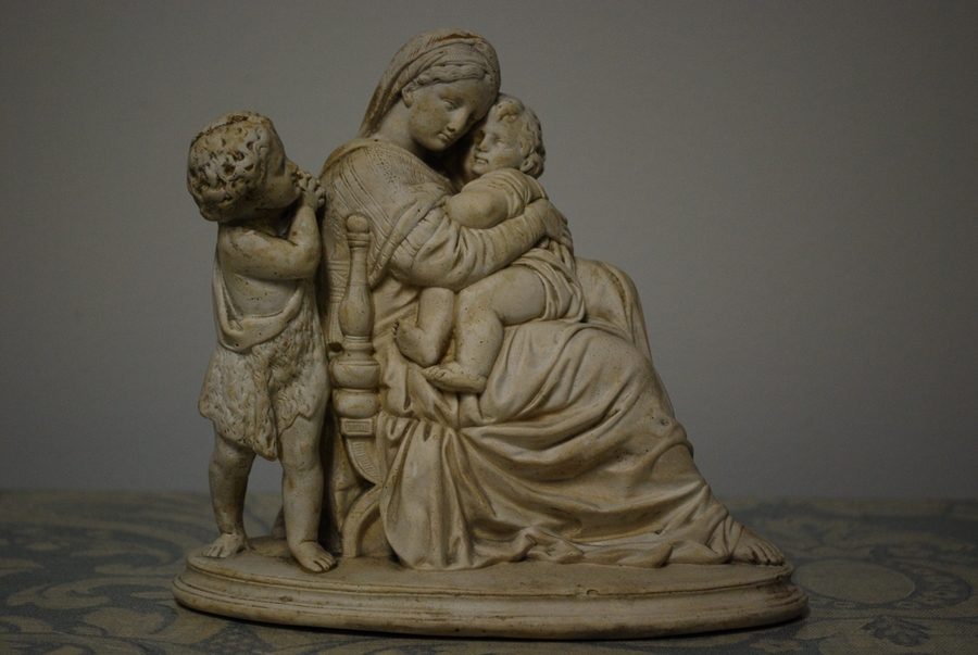 An off-white, plaster sculptural group depicts an idyllic scene. A small child peeking around the shoulder of a larger, seated woman cradling another chubby child close. She reproduces the chair form with her solid body and flowing robes.