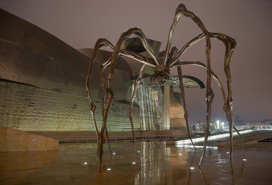 A monumental steel spider has eight slender, misshapen, and curving legs. The spider's body is suspended high in the air, and an egg sac is visible beneath the body. The sculpture is photographed beside a modern building at nighttime.
