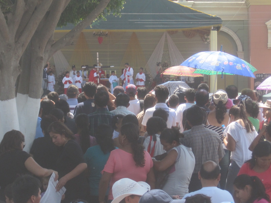 A congregation gathers in front of an outdoor tent and stage. A priest and altar servers gather on the stage around an altar and a large wooden cross.