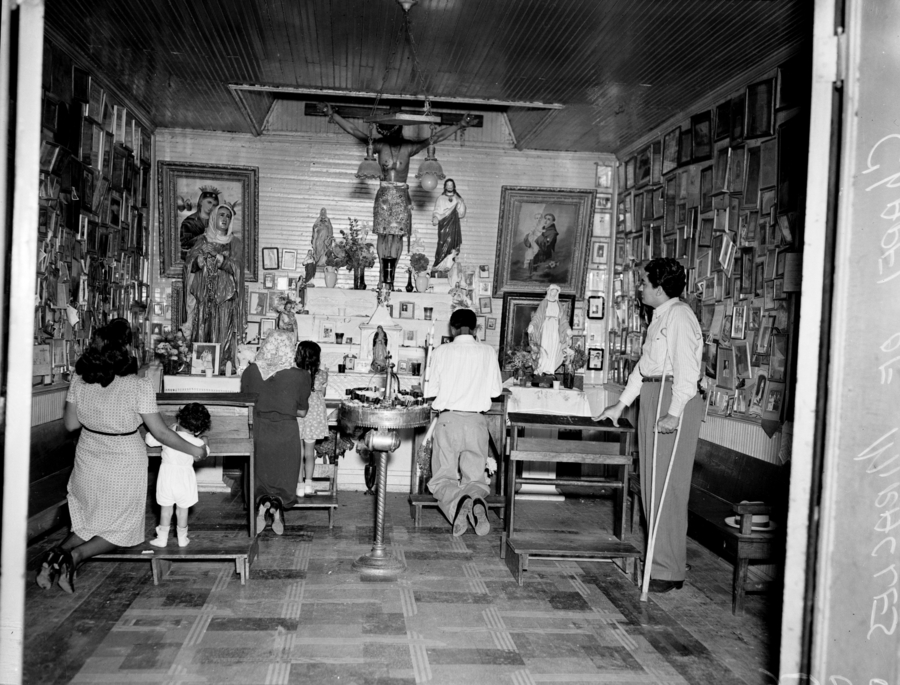 A black and white photo shows a space with adults and children on kneelers before an altar arrayed with devotional statues. The walls are lined with small framed images. One devotee stands on crutches. 