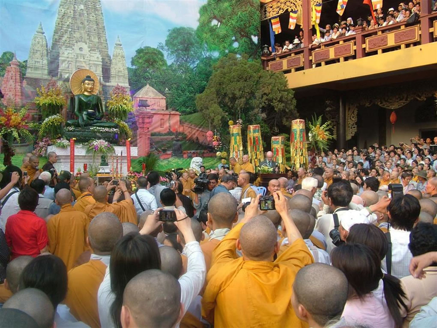 A mass of people gather around a jade sculpture of a seated Buddha. They stand on the ground and on building balconies nearby to take pictures of the sculpture. There is a printed backdrop hung behind the work.