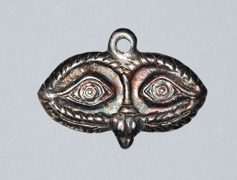 A metal, mask-shaped pendant depicts a pair of slightly raised, stylized eyes with circular pupils.