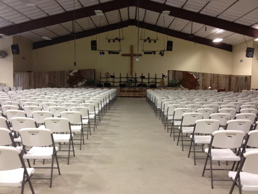 White folding chairs line an otherwise sparse assembly hall. A wooden platform stands at the far end of the hall and a cross hangs on the wall. Sound system wires hang down from the gabled ceiling.