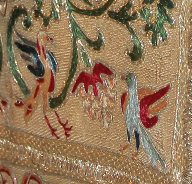 A detail of the embroidery on a gold miter shows two colorful birds gathered around a bushel of pale grapes.