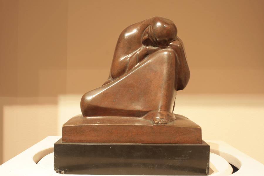 A view of a sculpture captures the triangle formed by the robed legs of a woman as she rests her head on her knee.