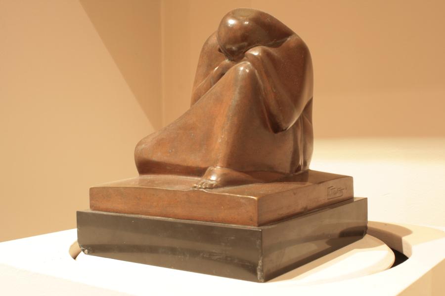 A woman lies her head on her knee in a shiny brown sculpture. Her body is abstracted beneath her robes, but a single foot peeks out from beneath the sculptured clothes.