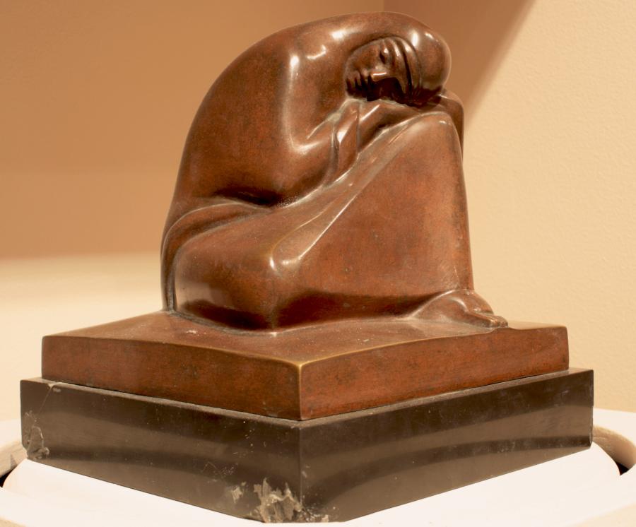 A brown stone sculpture depicts a woman sitting and resting her head on her right knee. Her body disappears beneath her robes. She forms a tight, closed mass. 