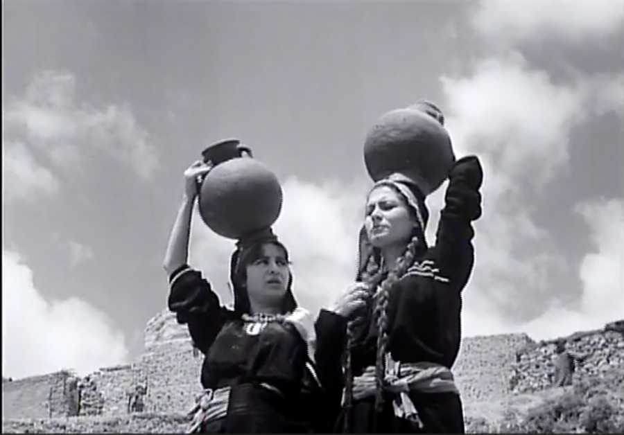 A black and white film still looks up at two women with handled jugs on their heads. The women wear dark tunics cinched at the waist, and they talk to each other while balancing the vessels..
