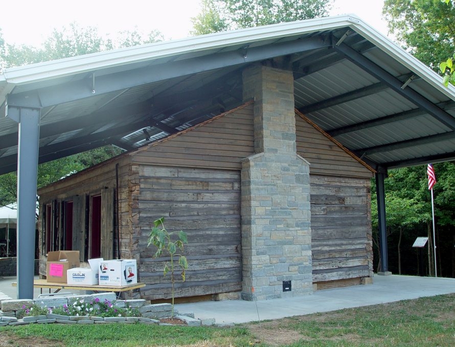 A photo captures a one-story wooden building with a gable roof and three entrance doors. The building stands beneath a modern metal awning. 