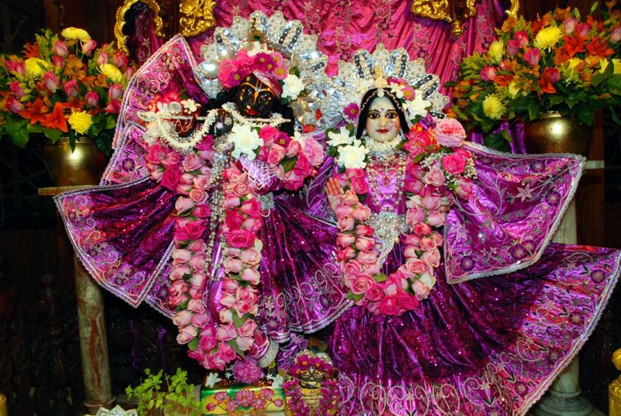 A pair of figures in bright pink dress and shiny white crowns wear rose garlands and flower crowns. The black-colored figure raises a flute to their mouth and the white-colored figure holds a right palm up and outward to the viewer. 