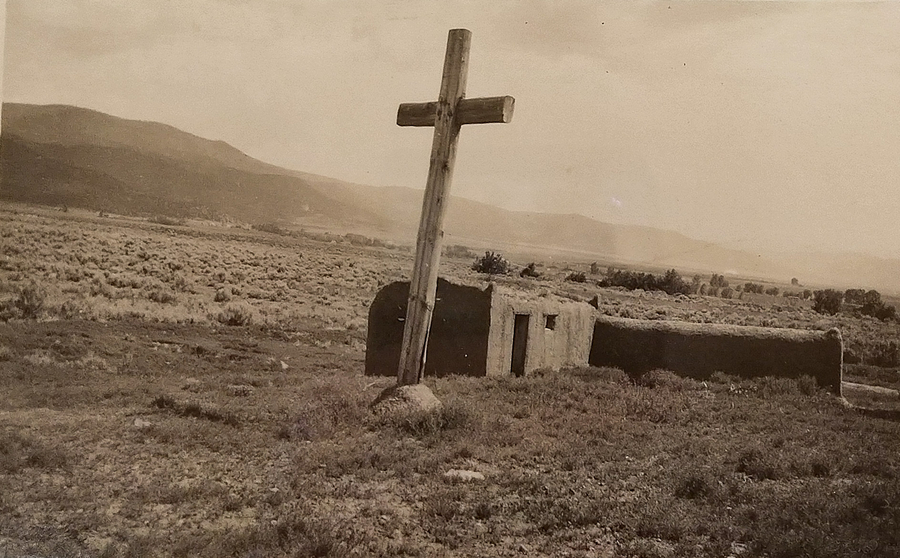 A wooden cross stands in a flat landscape in this sepia photograph. An adobe structure stands directly behind the cross and mountains lie in the background.