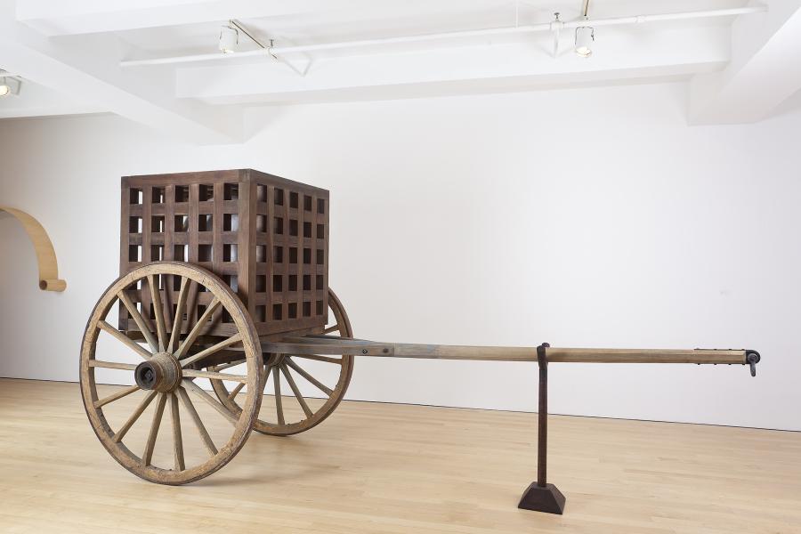 A large wooden cube sits atop a wooden cart. The cart is a platform with two large wheels and a long harness pole.