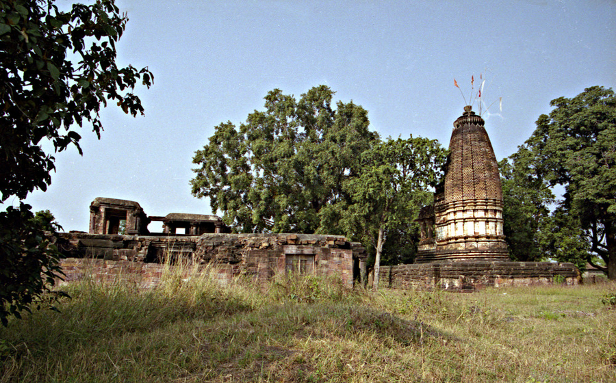 A stone temple complex stands in a grassy, tree-filled landscape. One section has been reduced to ruins but a stone building with a geometric, decorative surface still stands. It has an elongated cone shape.   
