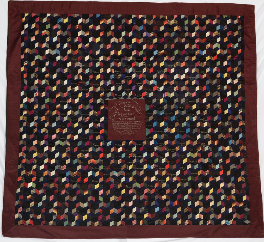 A dark-colored quilt has arrays of colorful, geometric cube shapes. Stitched in between the cubes are different names.
