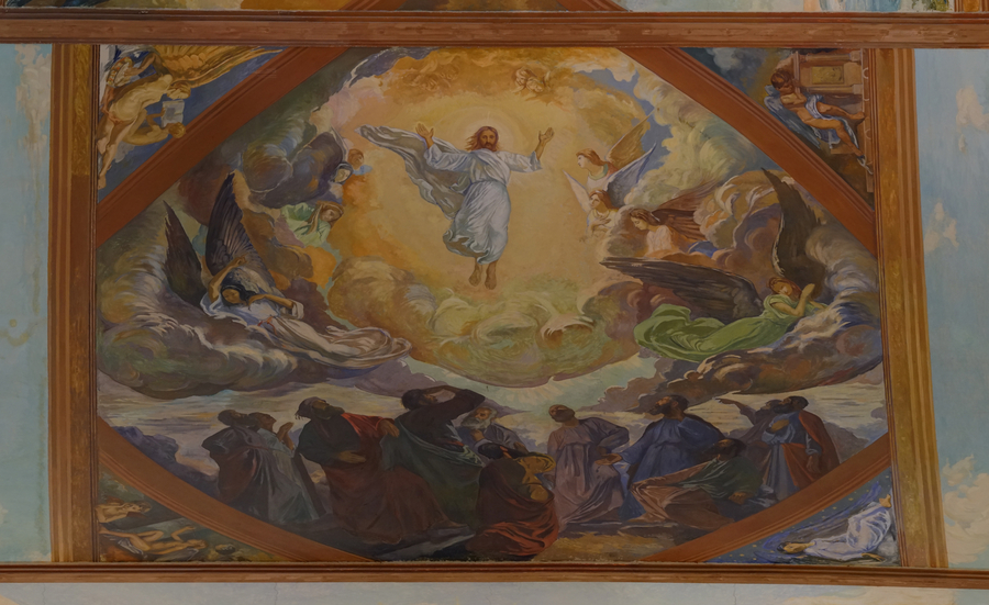 A section of a mural painting in a church depicts a Resurrection scene. Christ is shown hovering in a bright, angel-filled sky. He wears a white robe and holds his arms outstretched. Robed onlookers are shown on the ground.