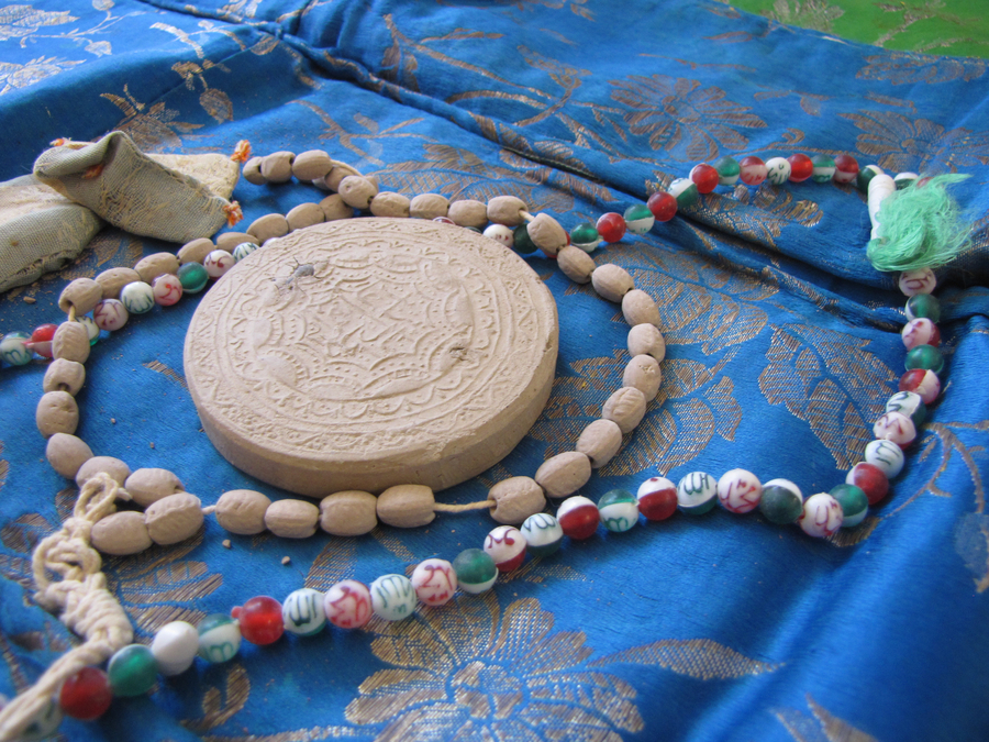 Devotional objects lie on a blue silken prayer rug. A circular, decoratively-carved piece of clay lies among two sets of prayer beads and two sachets.