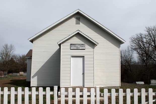 A one-story, wooden church with white siding stands behind a short wooden fence. A shorter chancel protrudes from the gable-end facade. A sign on the chancel reads, "BONDS CHAPEL METHODIST CHURCH."