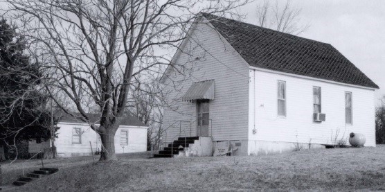 A black and white photo captures a one-story, gable-front church with a short rectangular nave. A simple door with a small awning stands at the entrance.