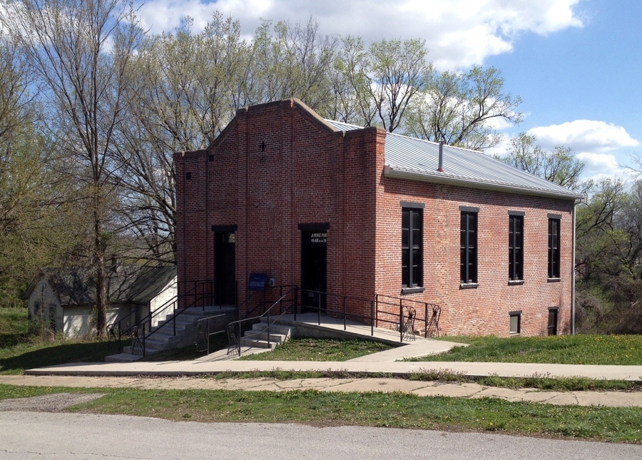 A brick building with two single-doors has a rectangular nave lined with windows. The facade features six low-relief brick pilasters and a simple, stepped gable.