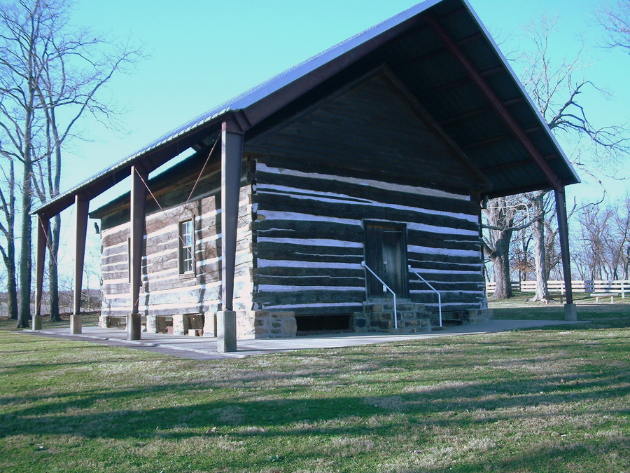 A log cabin style building has a gable roof. The sides have a striped brown and white appearance from rough wood planks. A steel awning stands above the building.