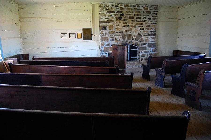 Two short rows of dark wooden pews stand within a small wooden room. They face a closed brick fireplace on the far wall.