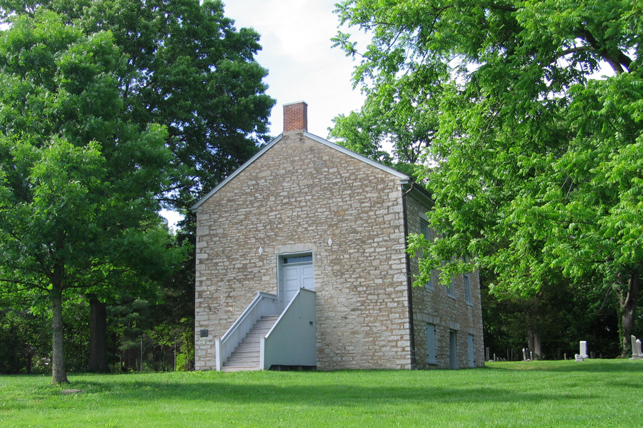 A three-storey, gable-end church is constructed of light colored stone. The front facade is plain and windowless. A set of steps leads up to a light blue door.