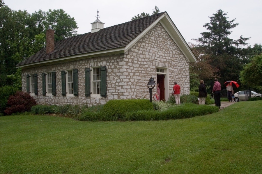 A small group of people gather outside a one-storey stone church. The building has a plain, windowless entrance and a gable roof. Four windows line the building's sides. 