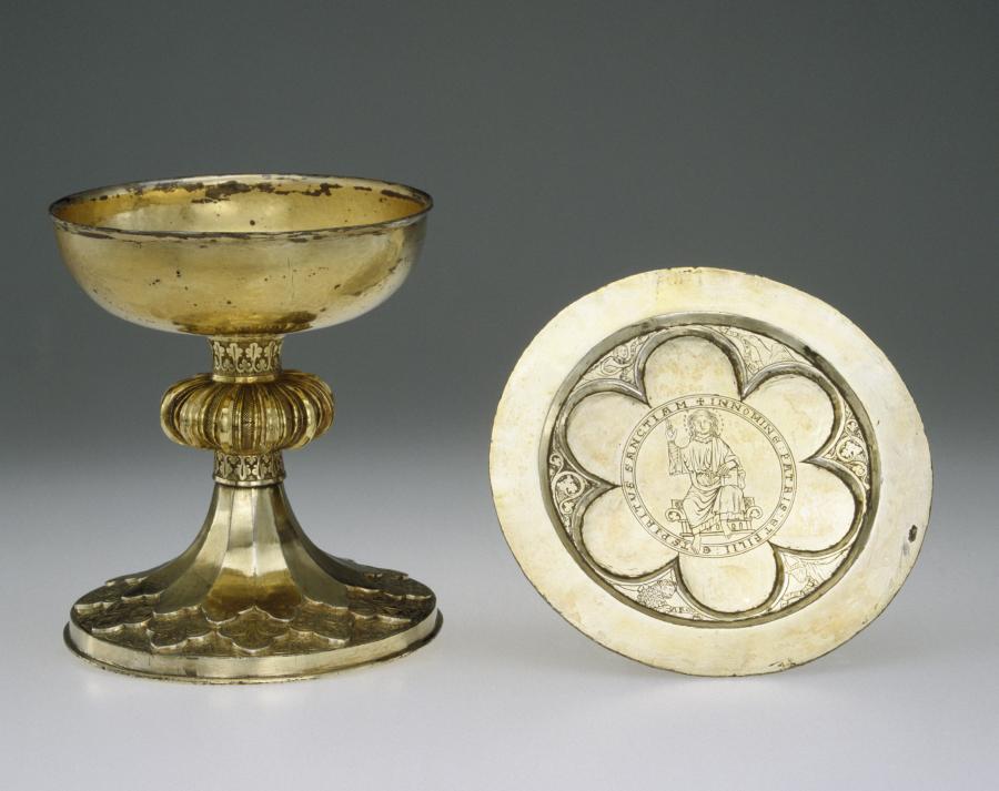 Two gold vessels are displayed side by side. The first is a footed cup with a ridged node and foliated base. A flat plate beside the cup has an engraving of an enthroned Christ. Christ is depicted in a six-lobed impression carved in the plate.