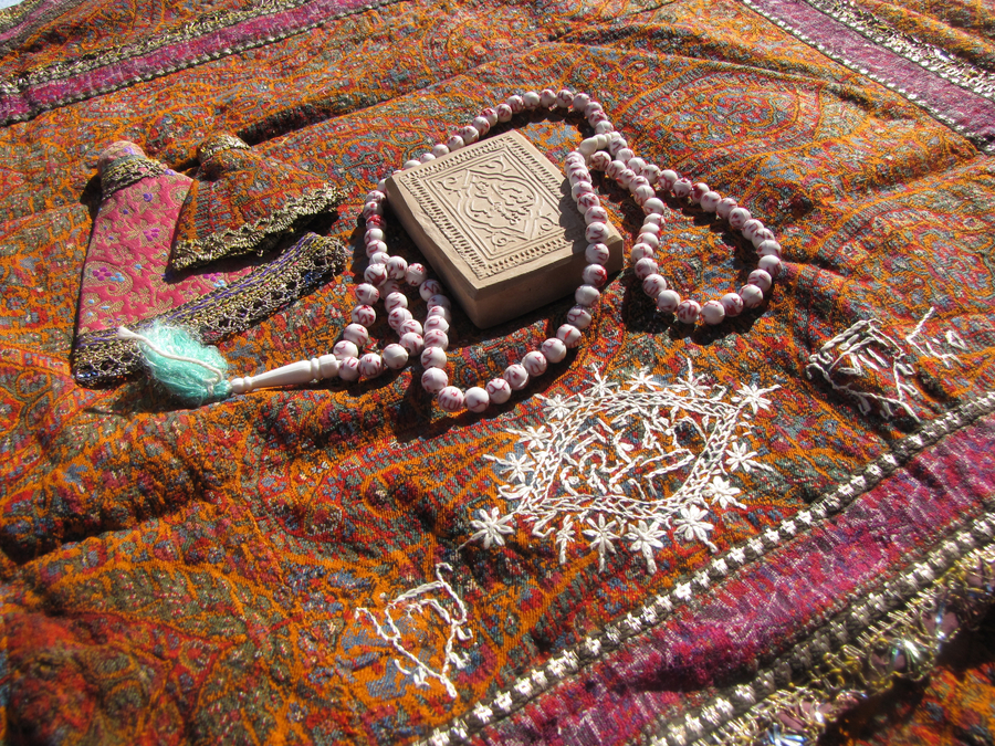 A patterened orange and pink prayer rug is scattered with devotional objects. A set of pink and white prayer beads lies around a square, decoratively-carved piece of clay.