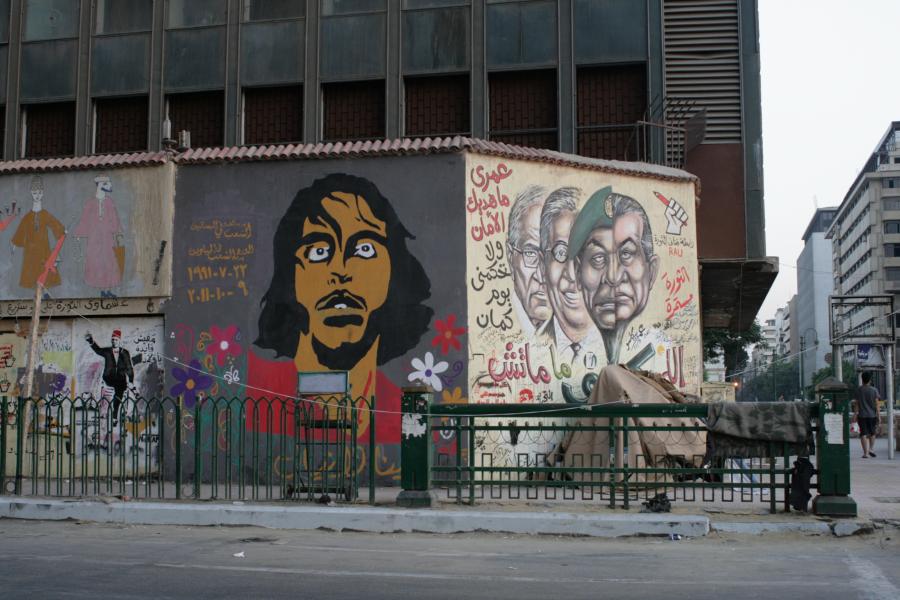 Mural and graffiti cover the walls of a squat city building. It includes a portrait of a tan-skinned man with dark hair and wide searching eyes. Another wall depicts a series of portraits of stern, older tan-skinned men.