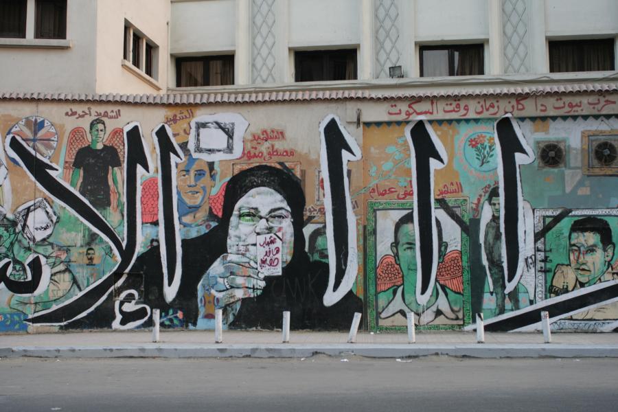 A colorful mural depicts a series of portraits on a building's horizontal wall. One large portrait depicts a man in a black robe holding up an inscribed card. Large black lettering is overlaid atop the figures.