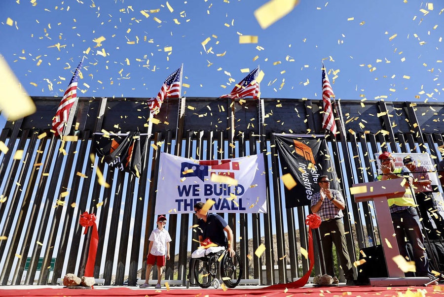 People stand and clap as confetti falls on a stage with a cut ribbon and sign that reads, "WE BUILD THE WALL." A young, light skinned boy and a man in a wheelchair are among the celebrants.
